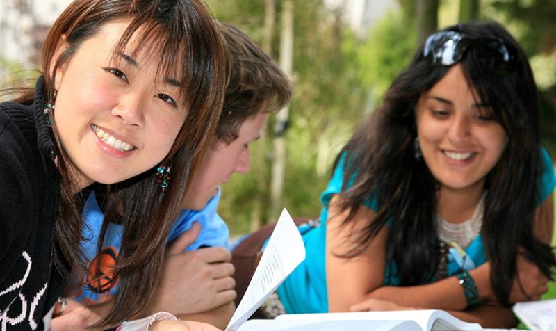 Three young women sit at a table, engaged in conversation while looking at a book in front of them.