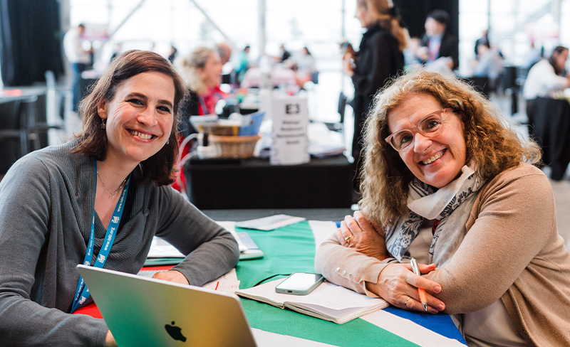 Two women engaged in discussion at a table, with a laptop open between them.