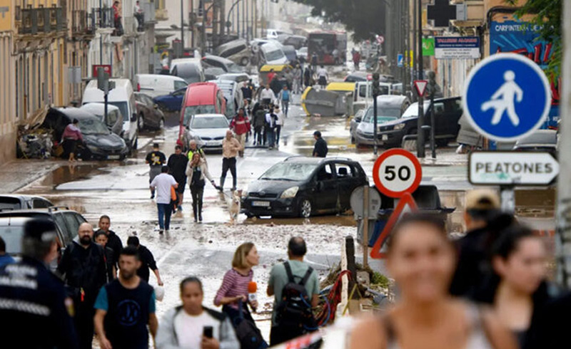 Pessoas navegam numa rua inundada da cidade, com água a cobrir o pavimento e edifícios visíveis ao fundo.