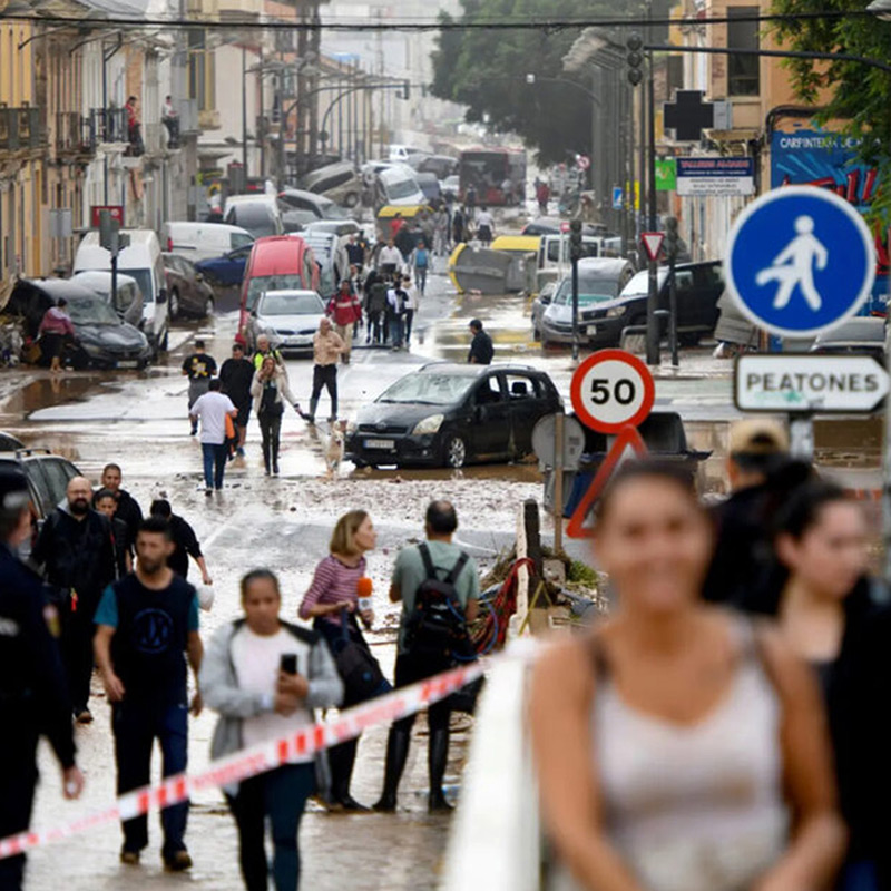 Pessoas navegam numa rua inundada da cidade, com água a cobrir o pavimento e edifícios visíveis ao fundo.